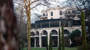 Un edificio con fachada de vidrio moderno, ventanas en arco y un patio ajardinado con setos podados y un sendero.