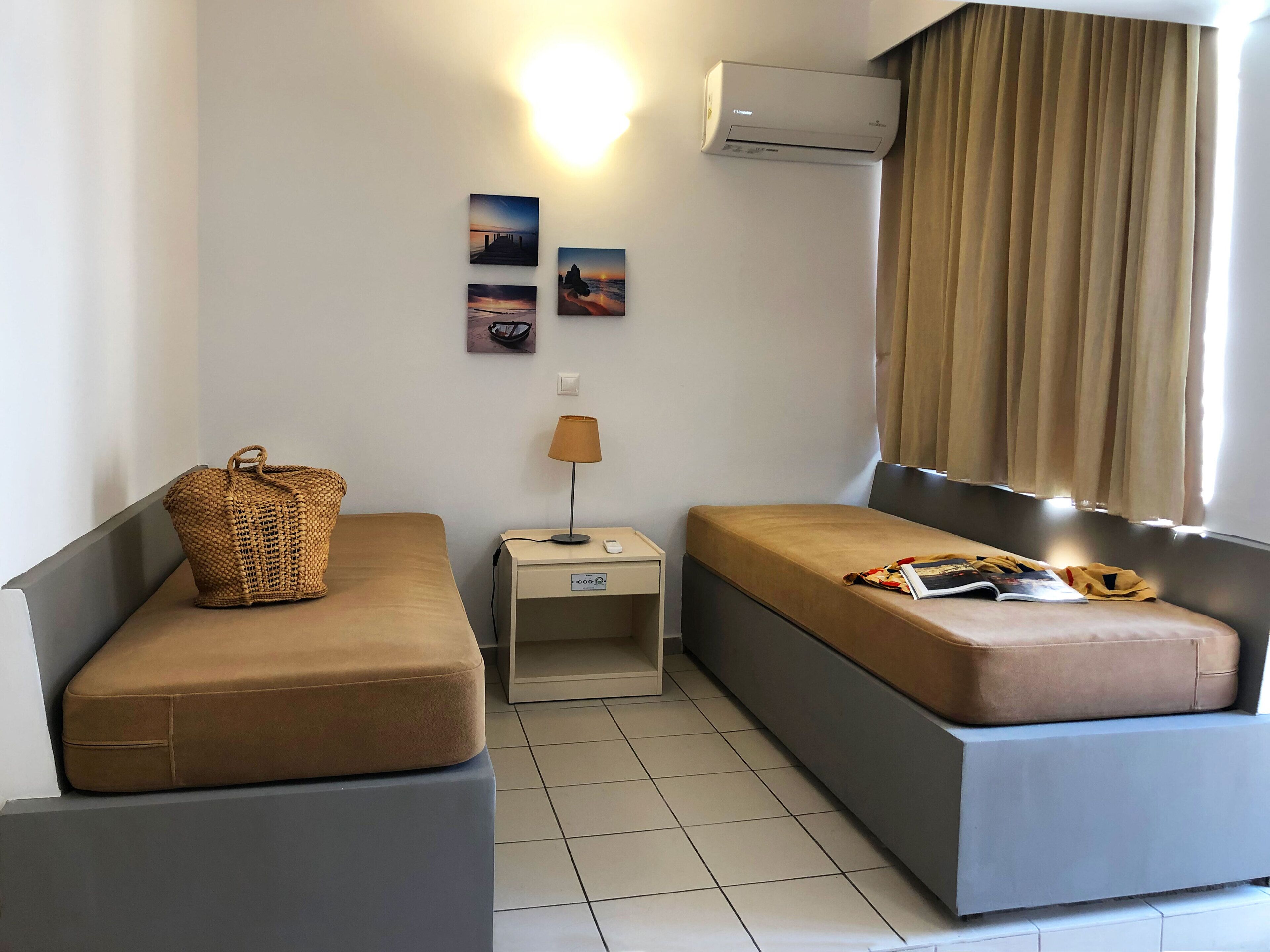 A small, modern room with two single beds, a bedside table with a lamp, a wall-mounted air conditioner, and a window with curtains.