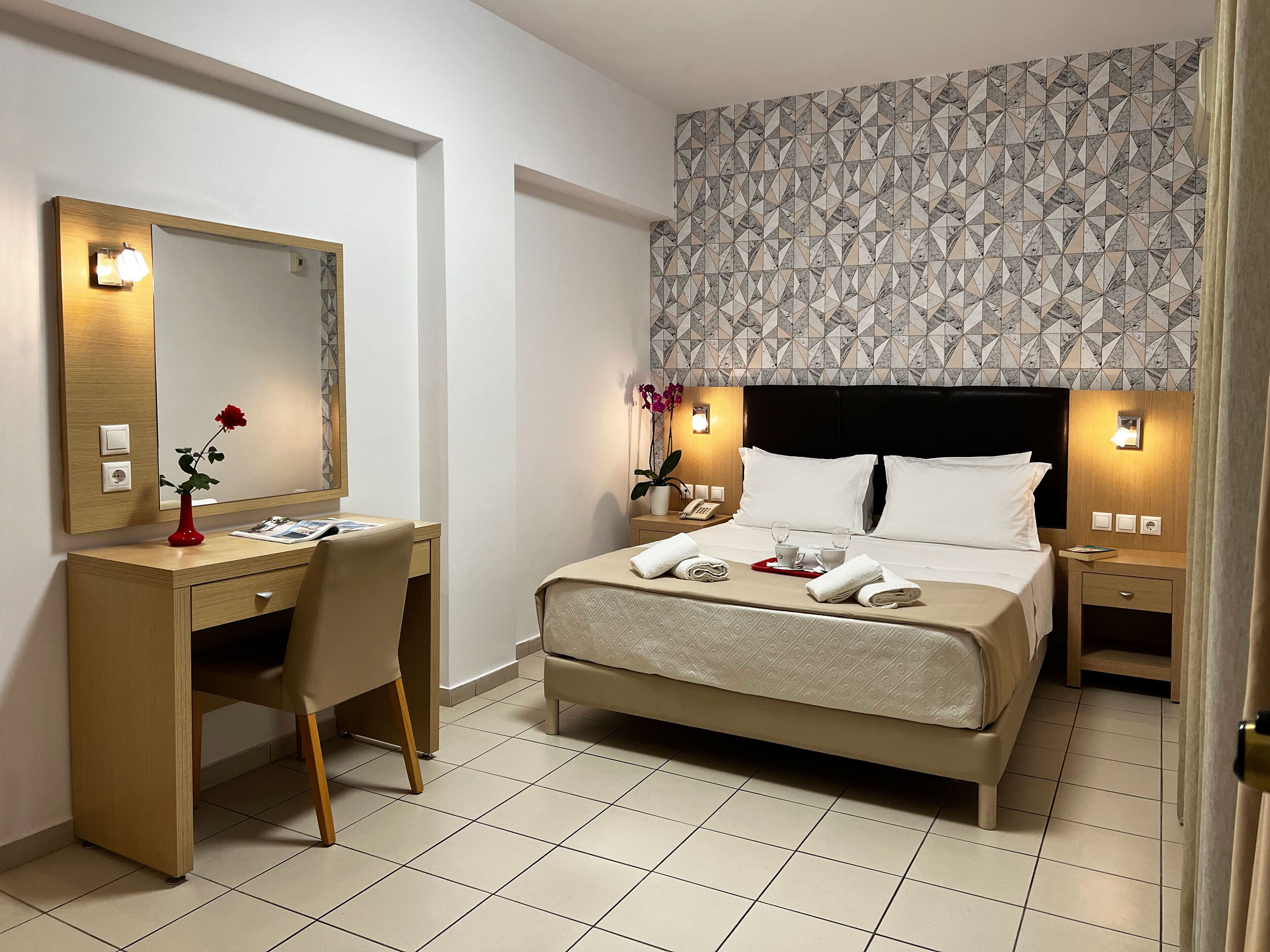 A hotel room with a bed, bedside tables, a desk, a chair, a mirror, and a patterned wall.