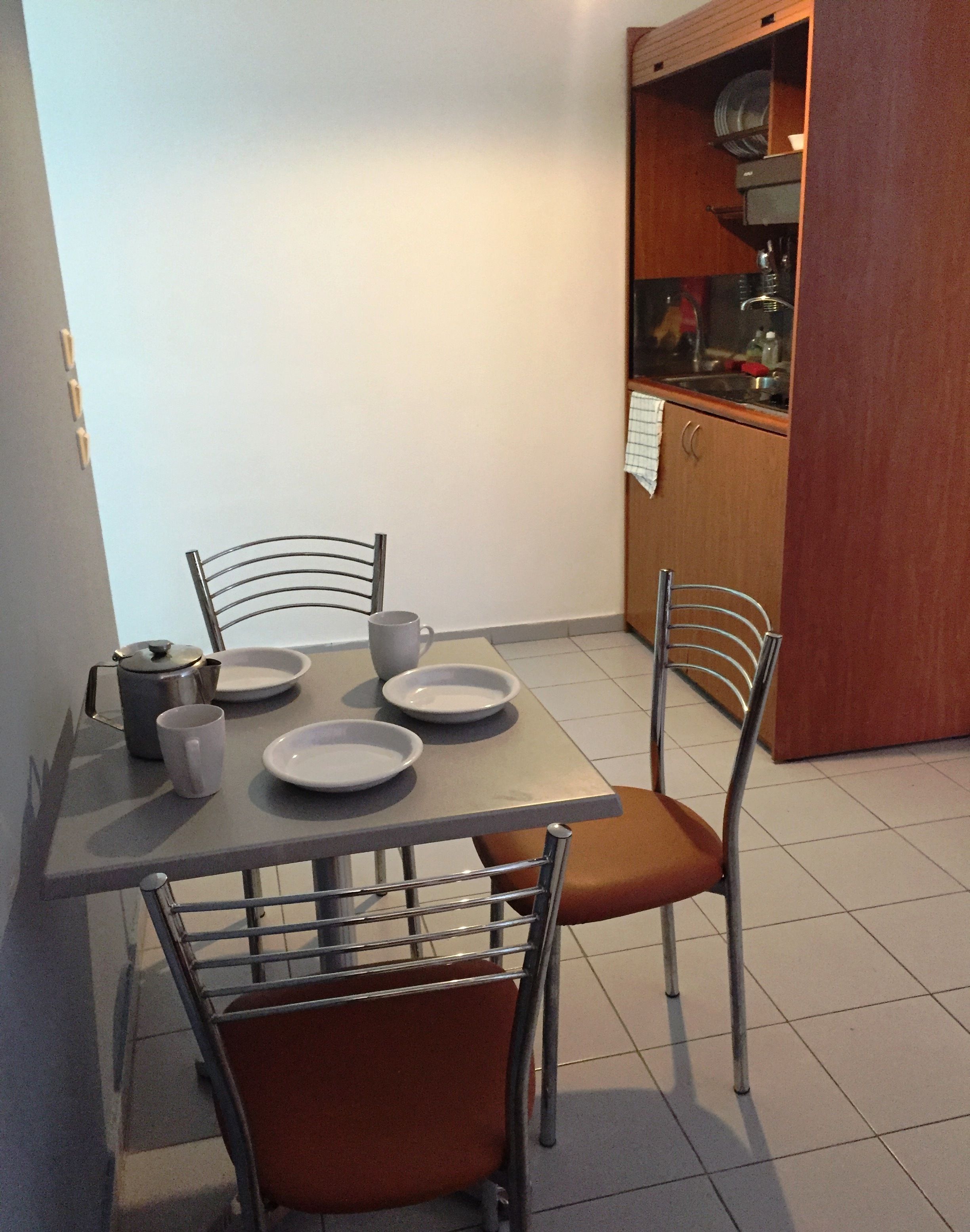A dining area with a table set for four, chairs, a kettle, and a cabinet with dishes.