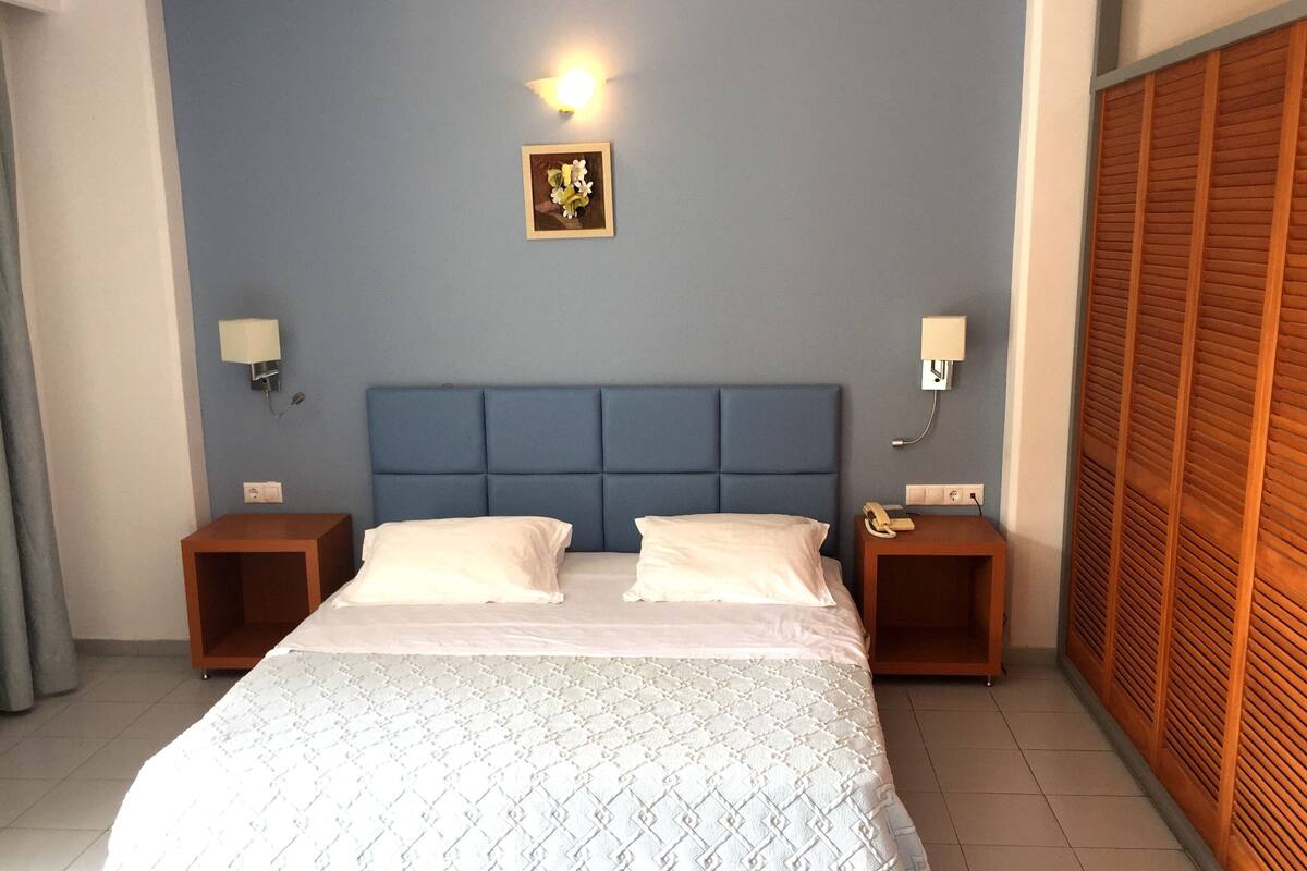 Een slaapkamer met een blauw hoofdeinde, twee nachtkastjes met lampen, een bed met een bed met een patroon en een houten garderobe.