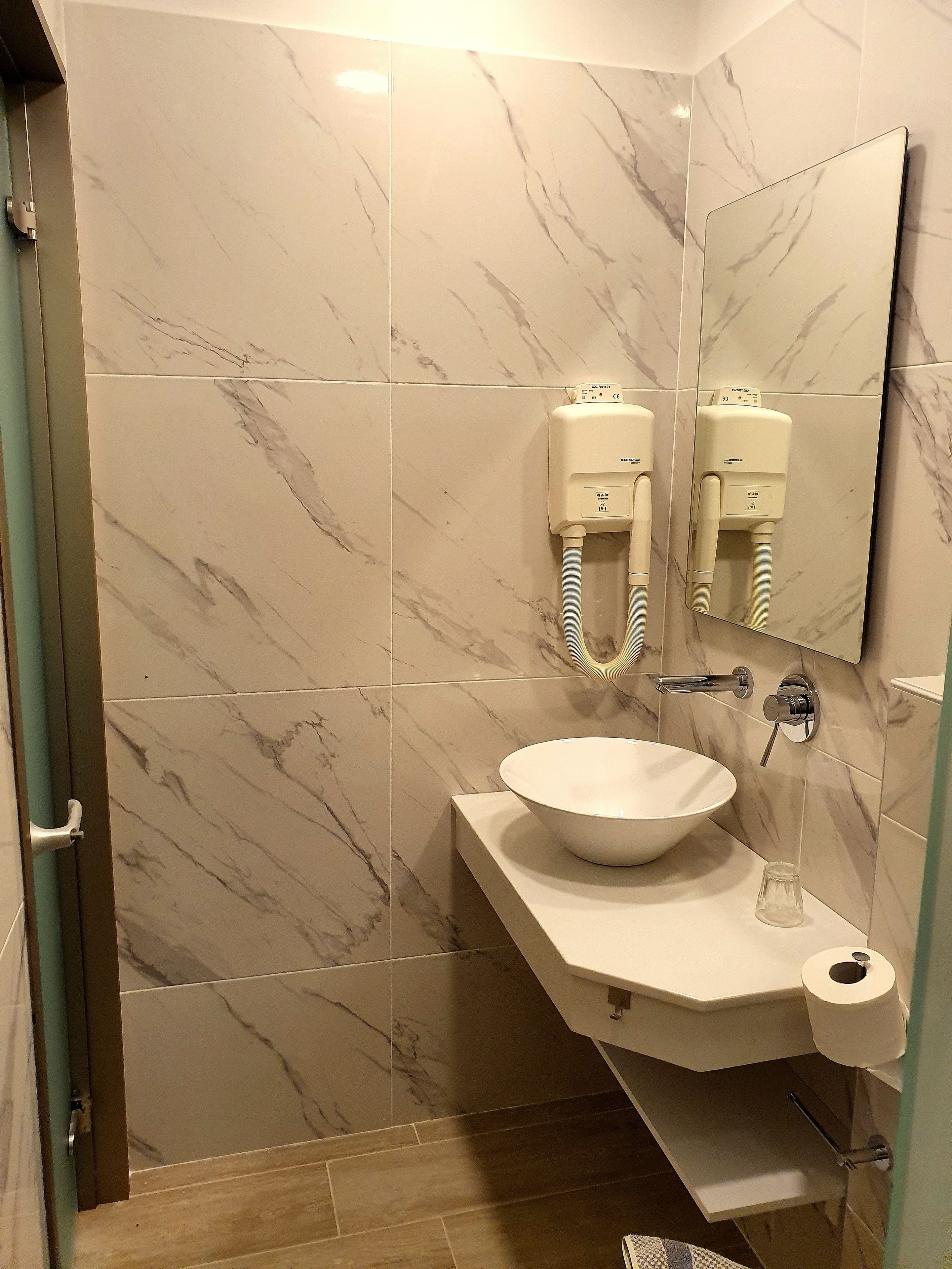 A bathroom with a marble-tiled wall, a white sink, a mirror, a hand dryer, and a toilet paper holder.