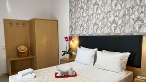Un lit bien fait, avec du linge de lit blanc, une table de chevet sur laquelle se trouve un plateau avec des tasses et des soucoupes blanches, une armoire en bois et un mur orné d’un motif.