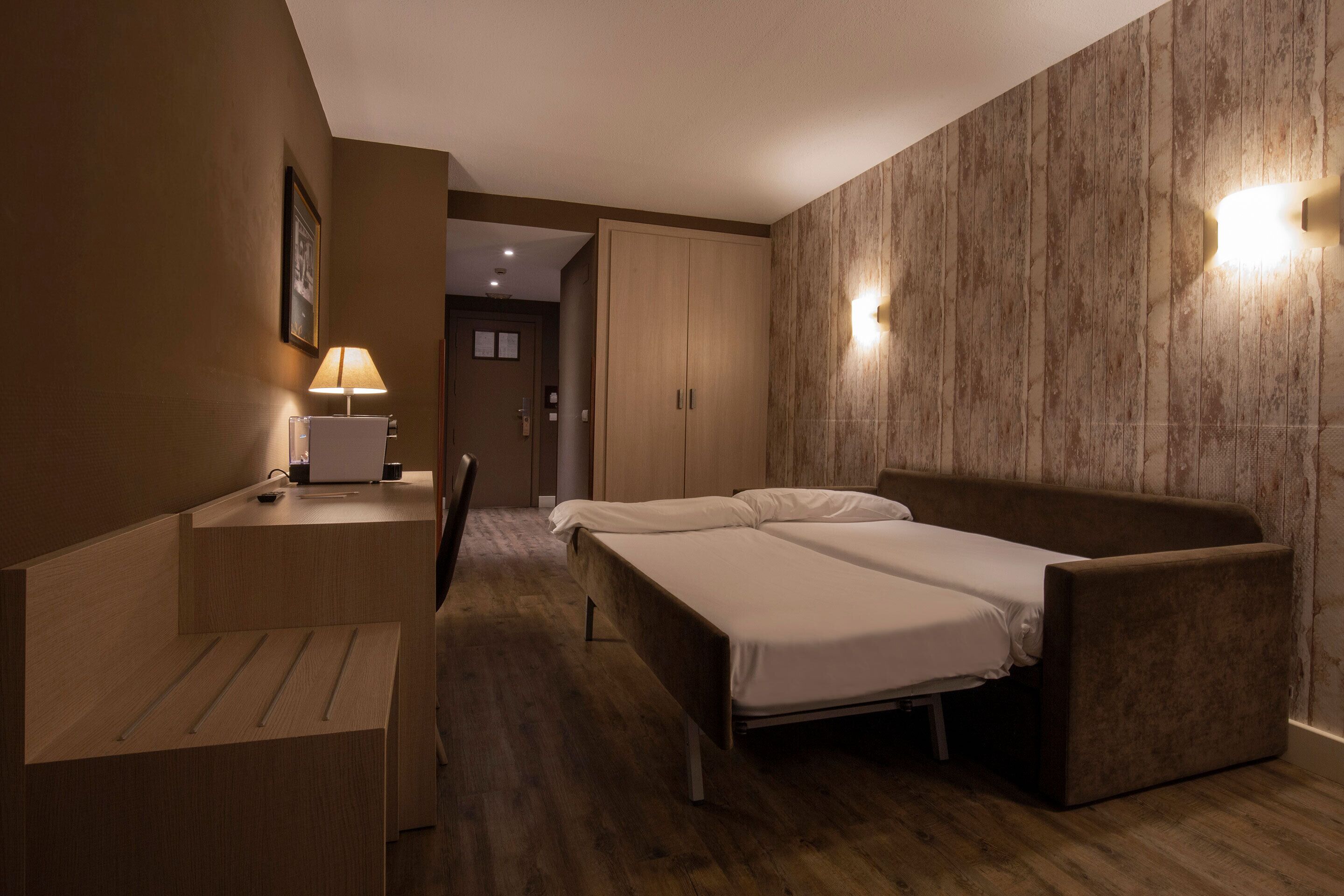 A hotel room with a bed, a desk, and a chair. There is a lamp on the desk and a picture on the wall.