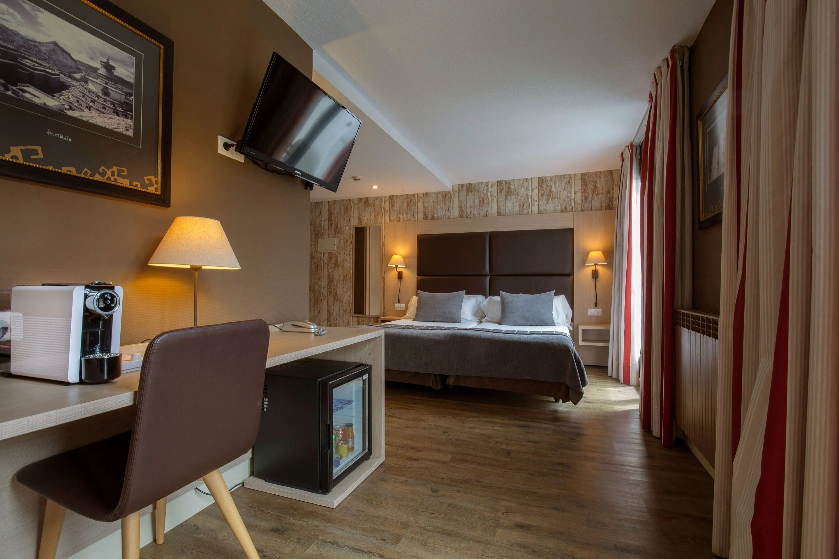 A hotel room with a bed, desk, chair, refrigerator, television, and a wall-mounted picture.