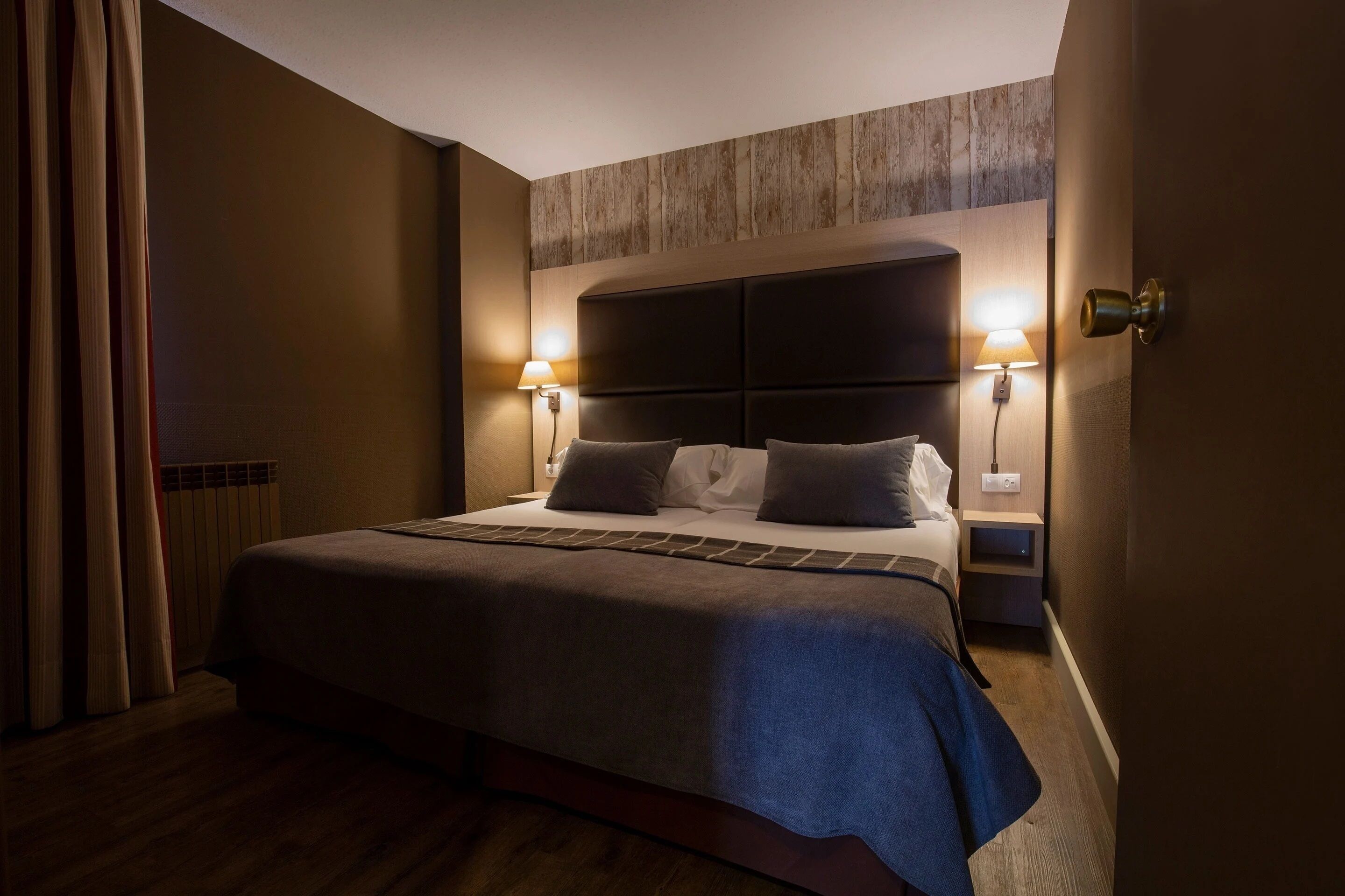 A hotel room with a large bed, two bedside lamps, a wooden headboard, and a nightstand.
