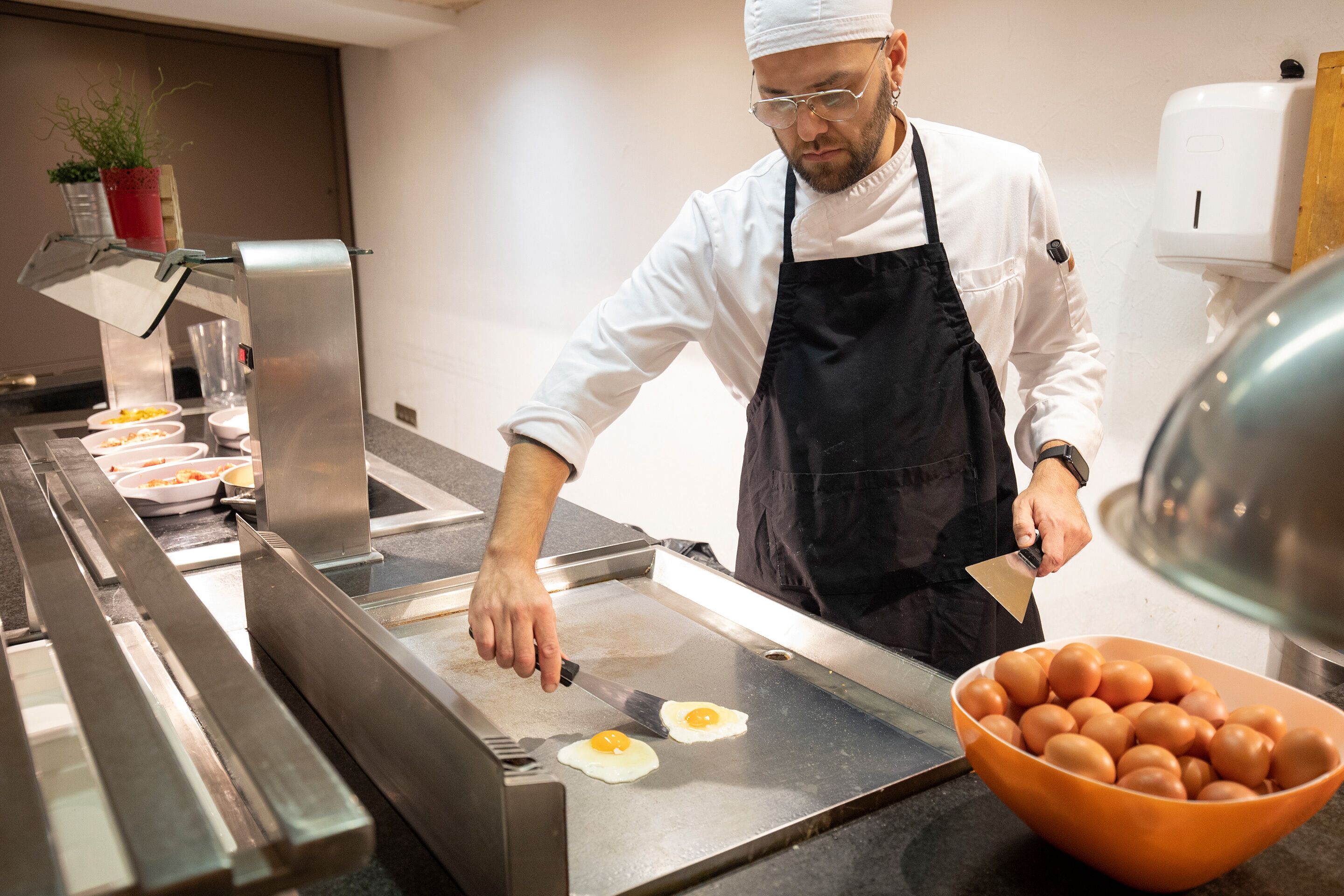 Un chef préparant des aliments dans une cuisine, avec une plaque de cuisson, un bol d’œufs et un distributeur d’eau.