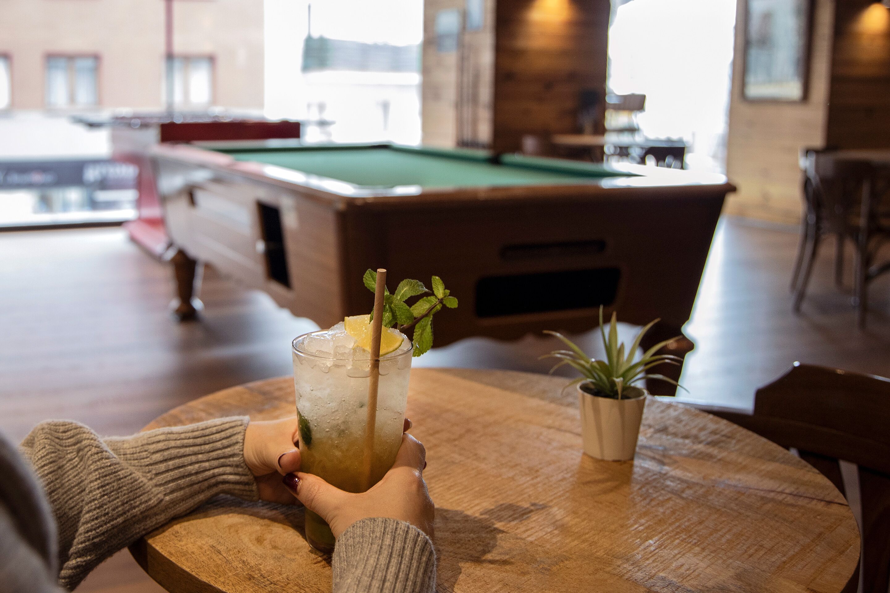 En person som håller i ett glas med en drink, en sugrör och en citronskiva, och sitter vid ett träbord med ett biljardbord i bakgrunden.