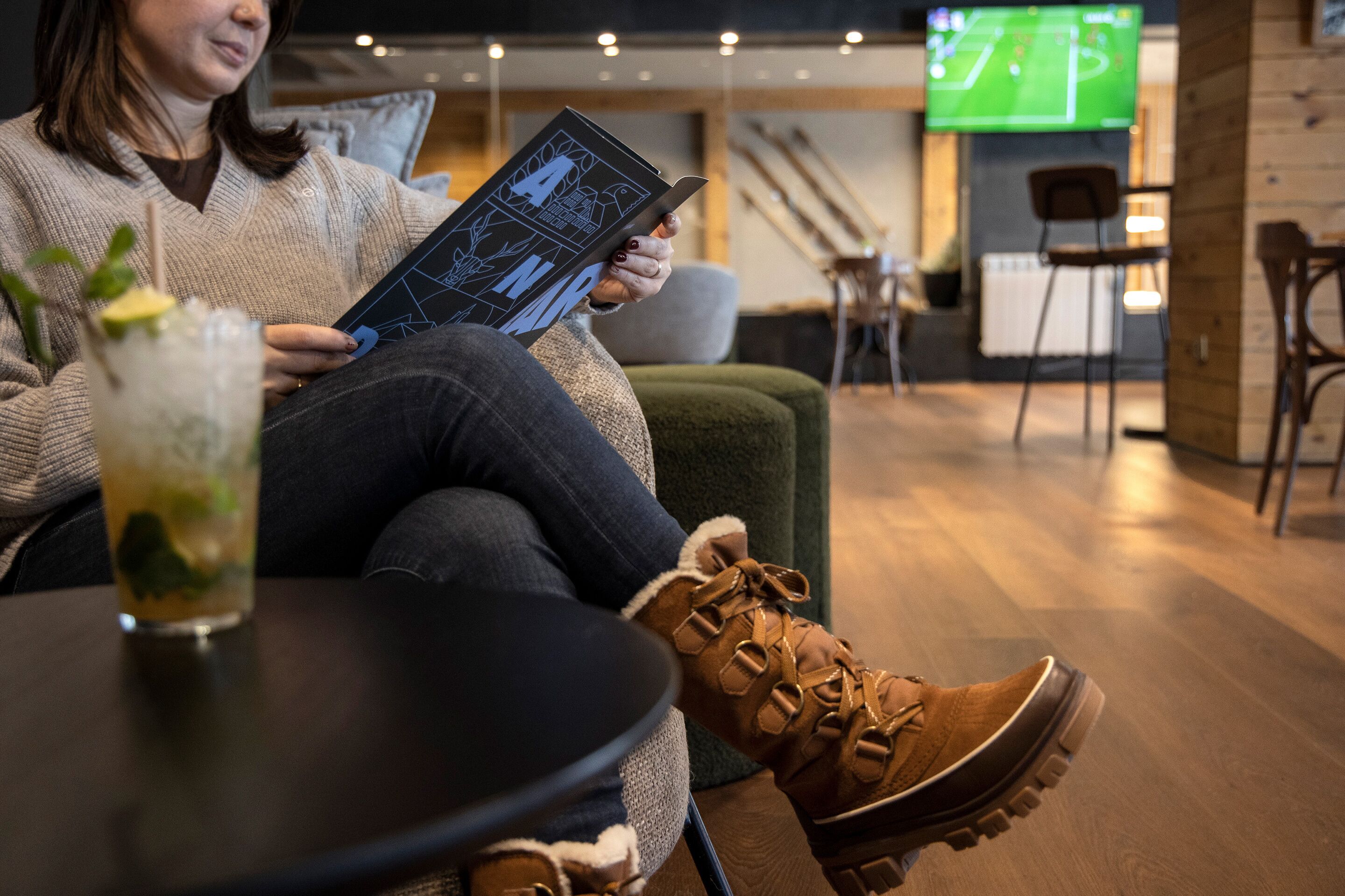 A person sitting on a couch reading a book, with a glass of drink on a table in front.