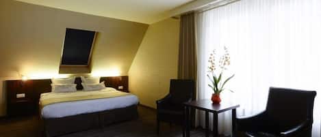 Habitación de hotel con cama, dos sillas, una mesita, una planta en maceta y un ventanal con cortinas.
