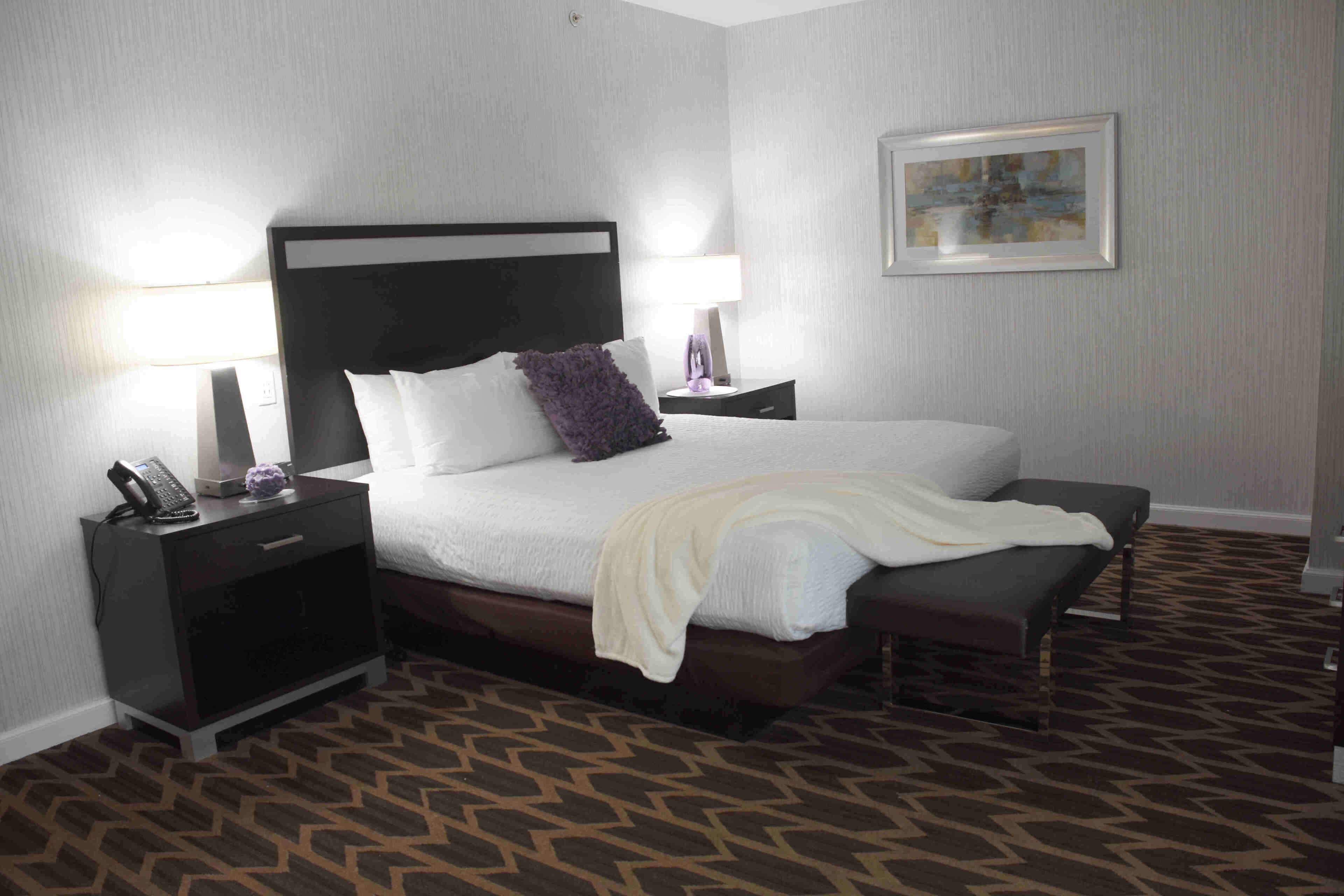 A hotel room with a large bed, two bedside tables, a nightstand, a bench, a lamp, and a framed picture on the wall.
