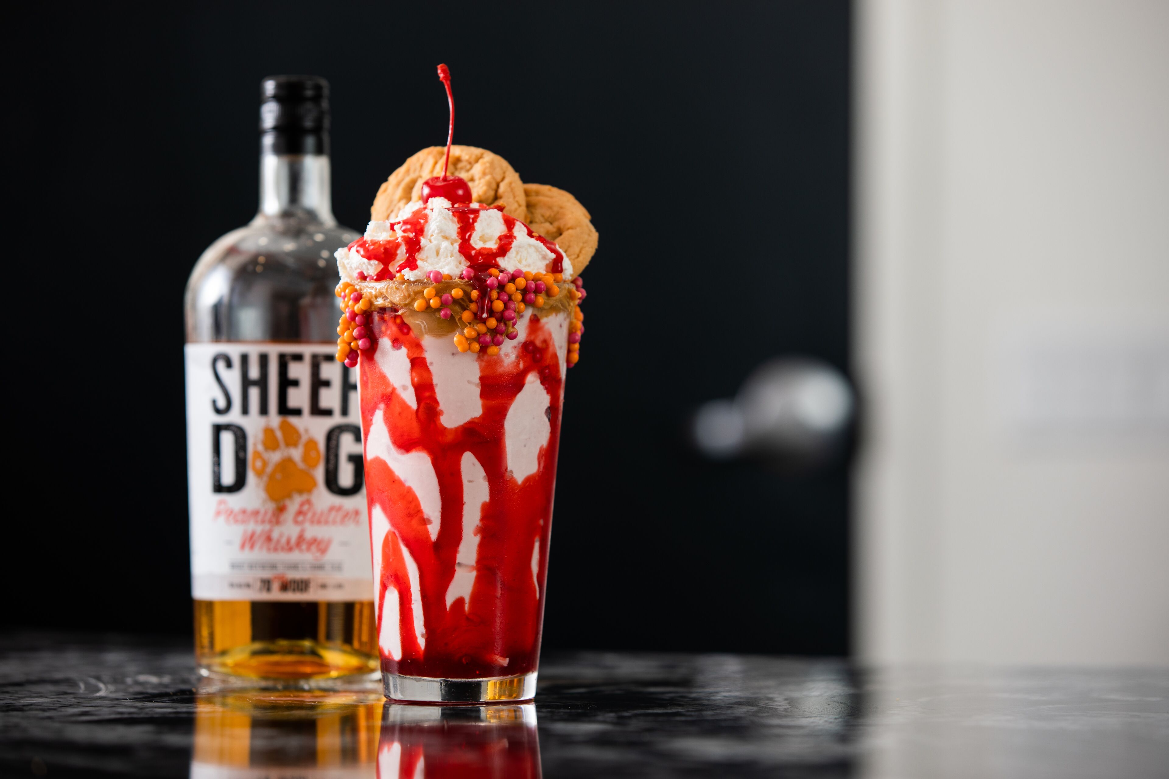 A dessert drink with a cookie on top, a cherry, and colorful sprinkles, accompanied by a bottle of whiskey.