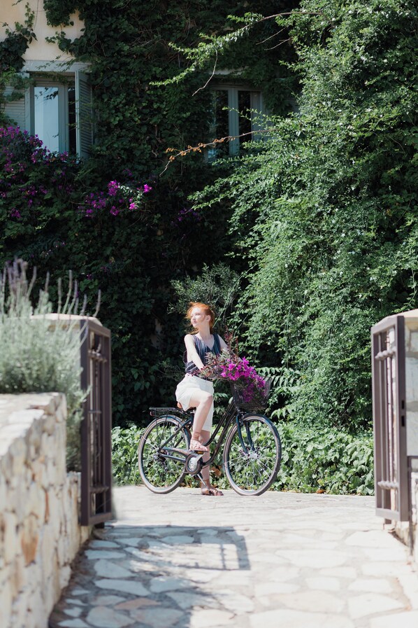 A person sitting on a bicycle in a garden area with a stone pathway, surrounded by lush greenery and a building with shutters. - Emelisse Nature Resort (Kefalonia)