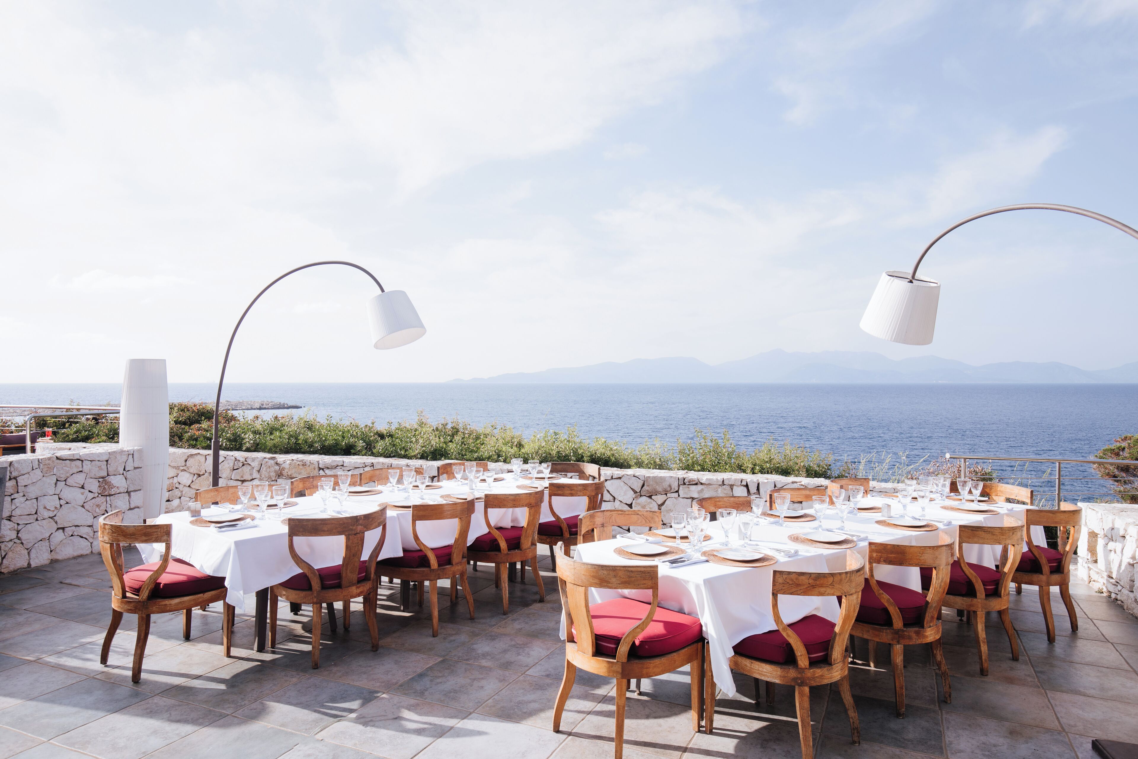 Un área de comedor al aire libre con mesas dispuestas para una comida, con vistas a un cuerpo de agua y montañas.
