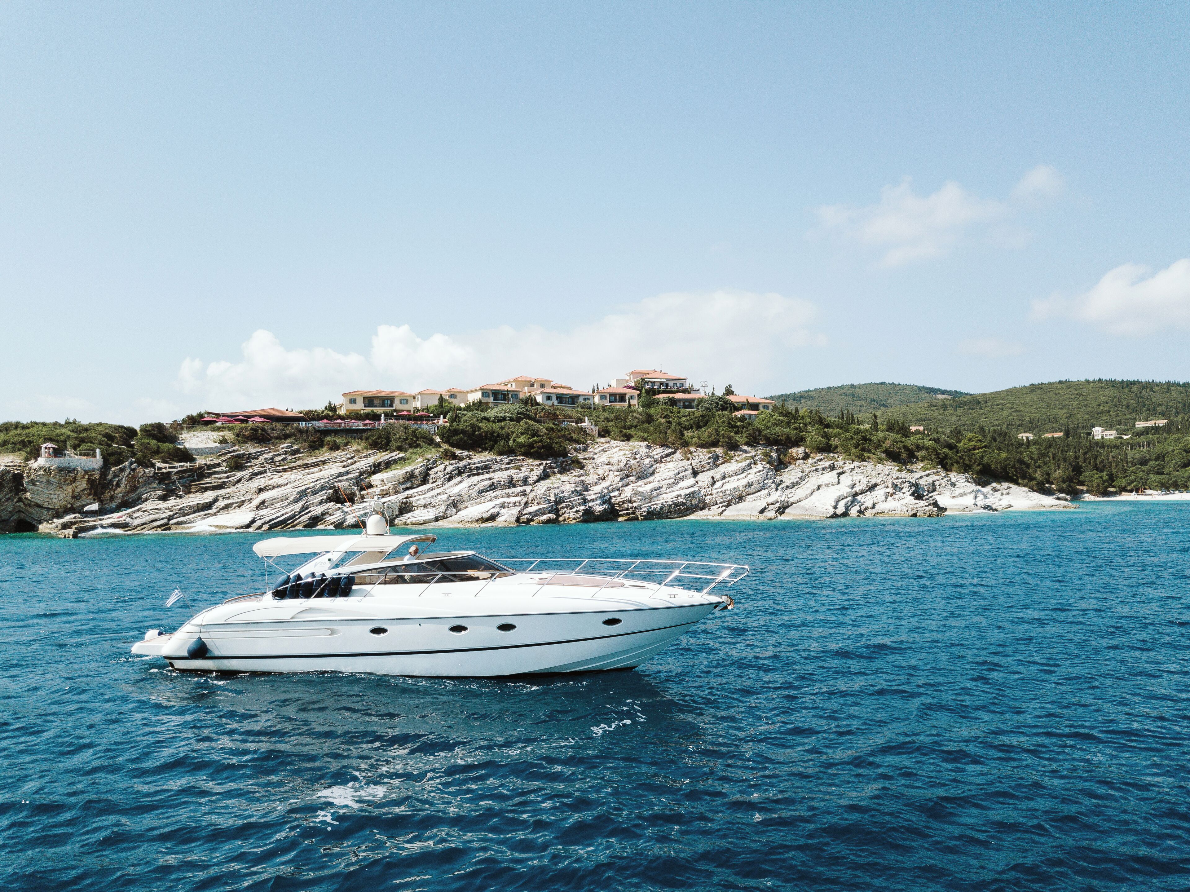 A yacht on a calm sea with a rocky coastline and houses in the background.