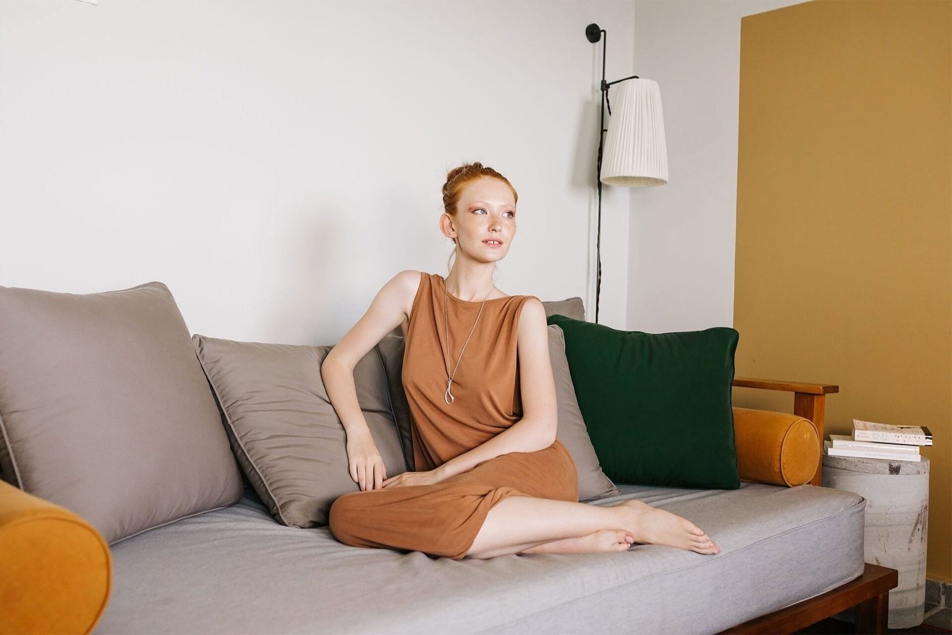 A person sitting on a grey sofa with multiple cushions, a floor lamp, and a stack of books on a side table.