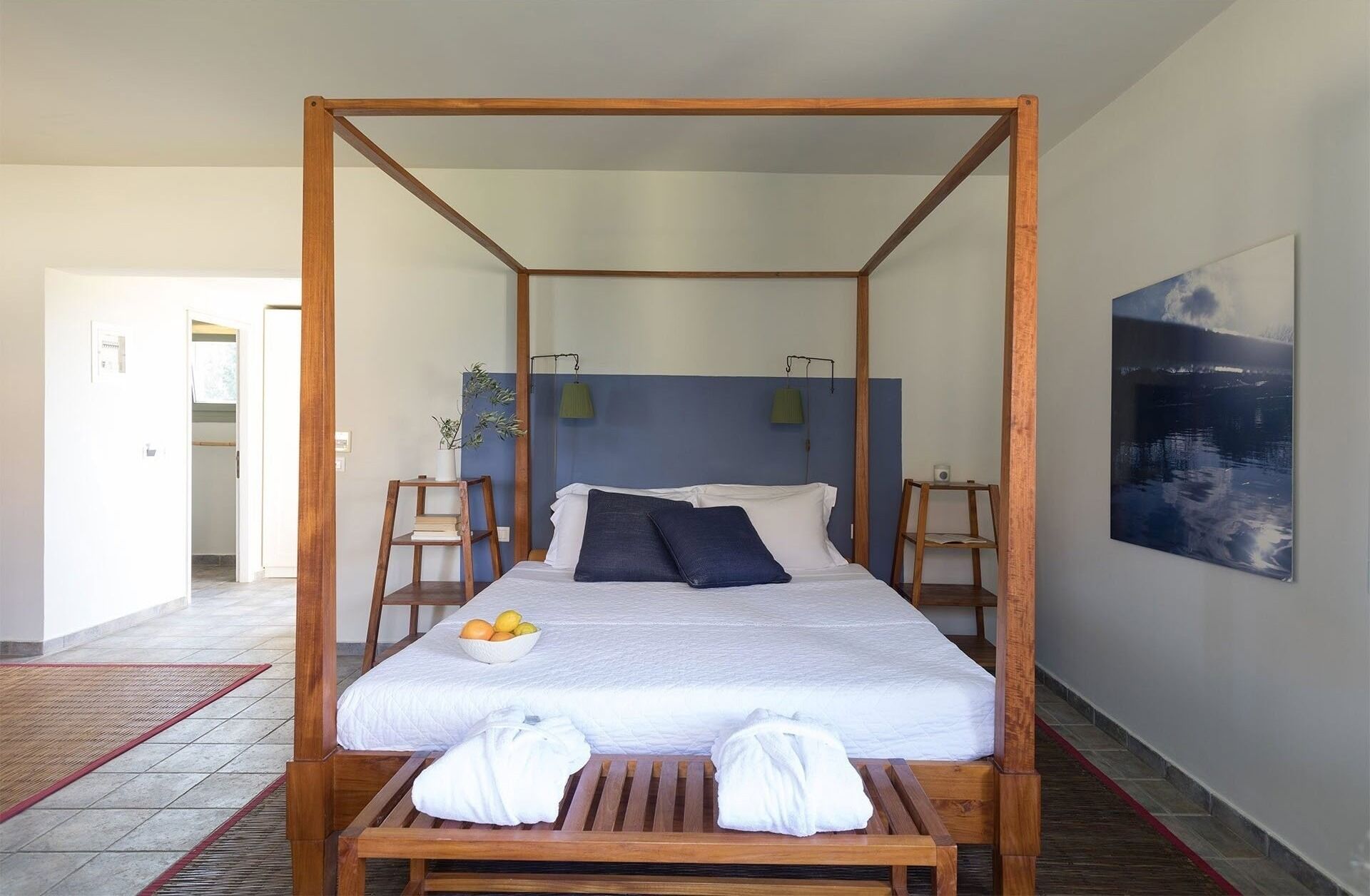 A four-poster bed with a white mattress, two pillows, a bowl of fruit, and two towels on a wooden bench.