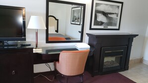 A hotel room with a desk, chair, TV, lamp, mirror, fireplace, and framed pictures on the wall. - Niagara Falls Courtside Inn (Niagara Falls)