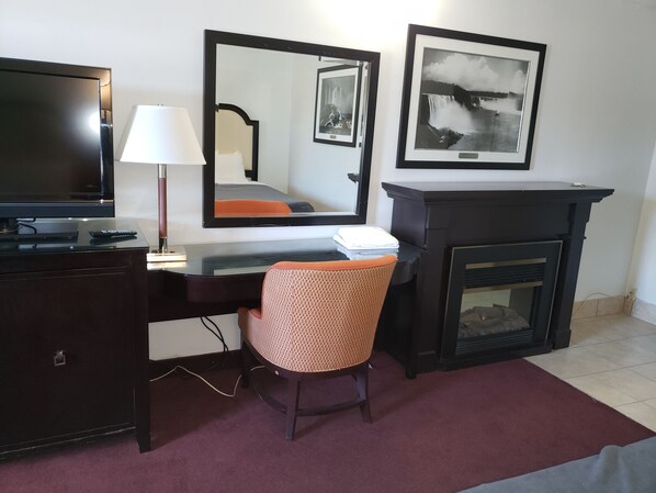 A hotel room with a desk, chair, TV, lamp, mirror, fireplace, and framed pictures on the wall. - Niagara Falls Courtside Inn (Niagara Falls)