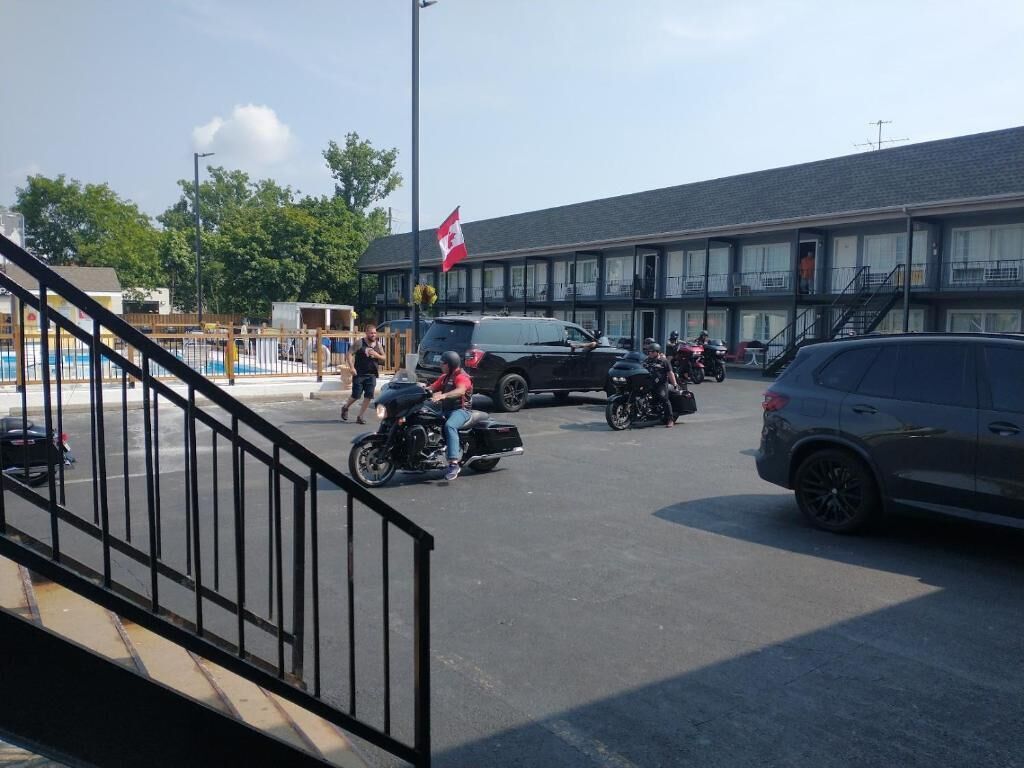 Un estacionamiento con varias motocicletas y automóviles, un edificio con una bandera y una escalera que lleva a una zona de piscina.