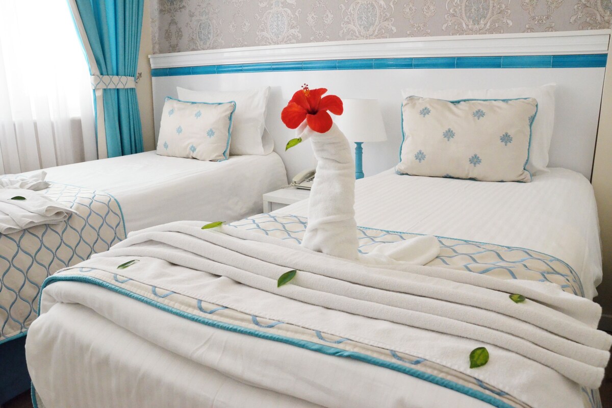 Habitación de hotel con dos camas, cada una con una colcha blanca y detalles azules, cabeceros a rayas azules y un papel tapiz con estampado floral.