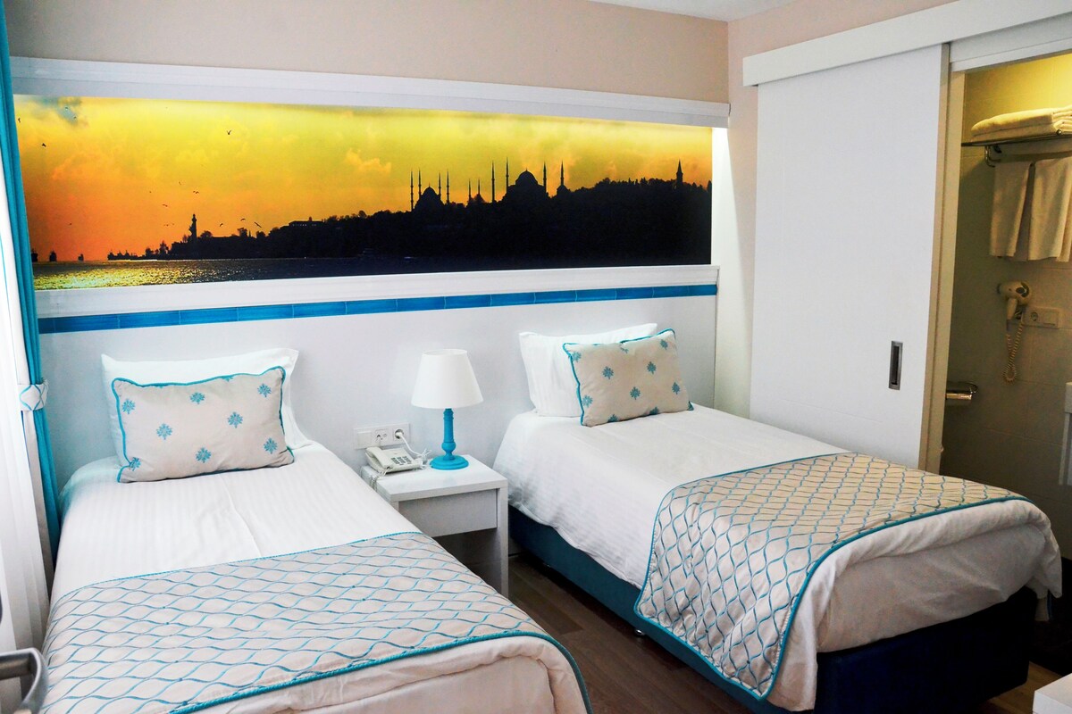 Habitación de hotel con dos camas, una mesita de noche con lámpara, un cuadro de un horizonte urbano y una puerta con perchero para toallas.