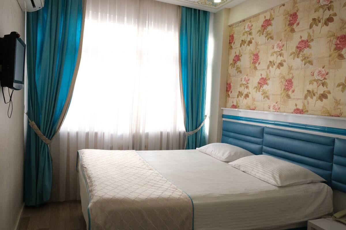 Une chambre d’hôtel avec un lit, des rideaux, un téléphone fixé au mur et un papier peint à motifs floraux.