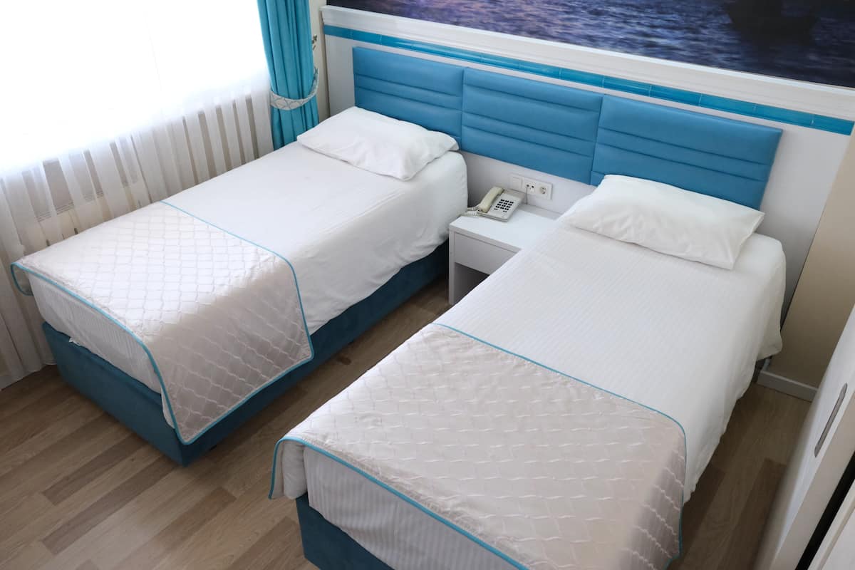 Une chambre d’hôtel avec deux lits, chacun doté d’une tête de lit bleue et de draps blancs. On y trouve une table de chevet avec un téléphone et une petite lampe.