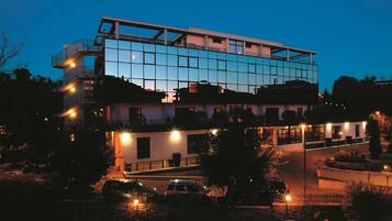Un edificio moderno de varios pisos con fachada de vidrio, varios balcones y un estacionamiento con automóviles.