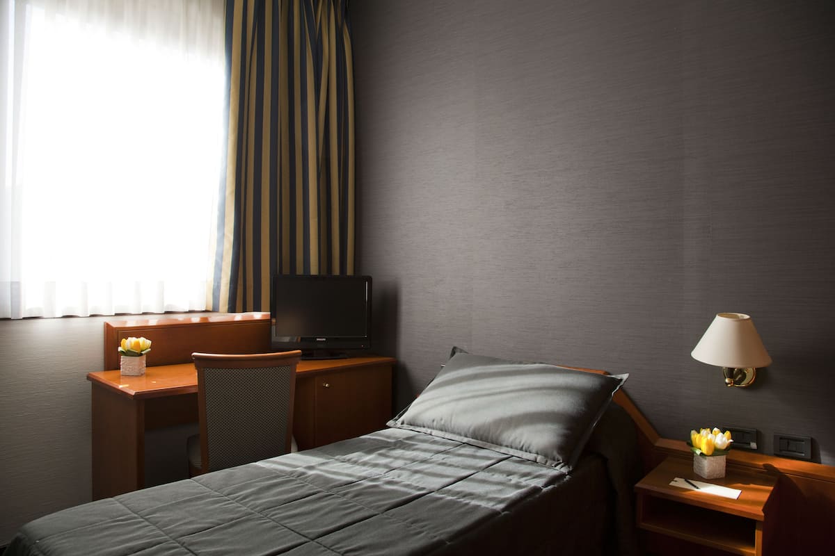 Hotellihuone, jossa on sänky, työpöytä tietokoneella, tuoli, televisio, lamppu ja ikkuna, jossa on verhot.