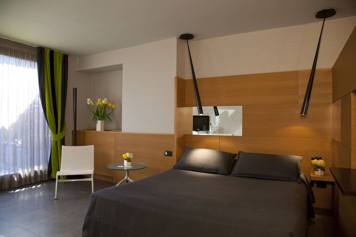 Moderni hotellihuone, jossa on suuri sänky, pieni pöytä maljakko täynnä kukkia, tuoli ja televisio, joka on sijoitettu puupaneeliseinälle.