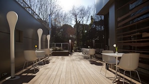 An outdoor patio with wooden flooring, white chairs, and tables, surrounded by a modern building with large windows and a railing.
