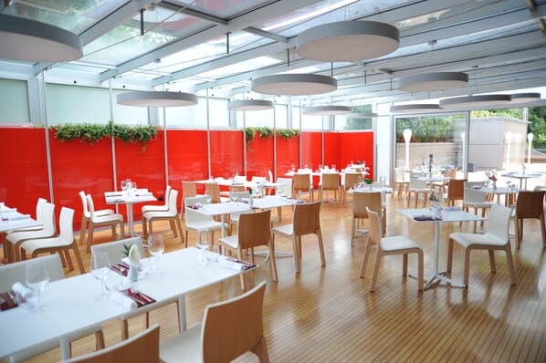Un restaurante moderno con mesas y sillas blancas, detalles en rojo y techo de vidrio.
