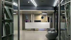 A modern reception area with a white reception desk, a round mirror, and two grey chairs. - Hotel Zone (Rome)