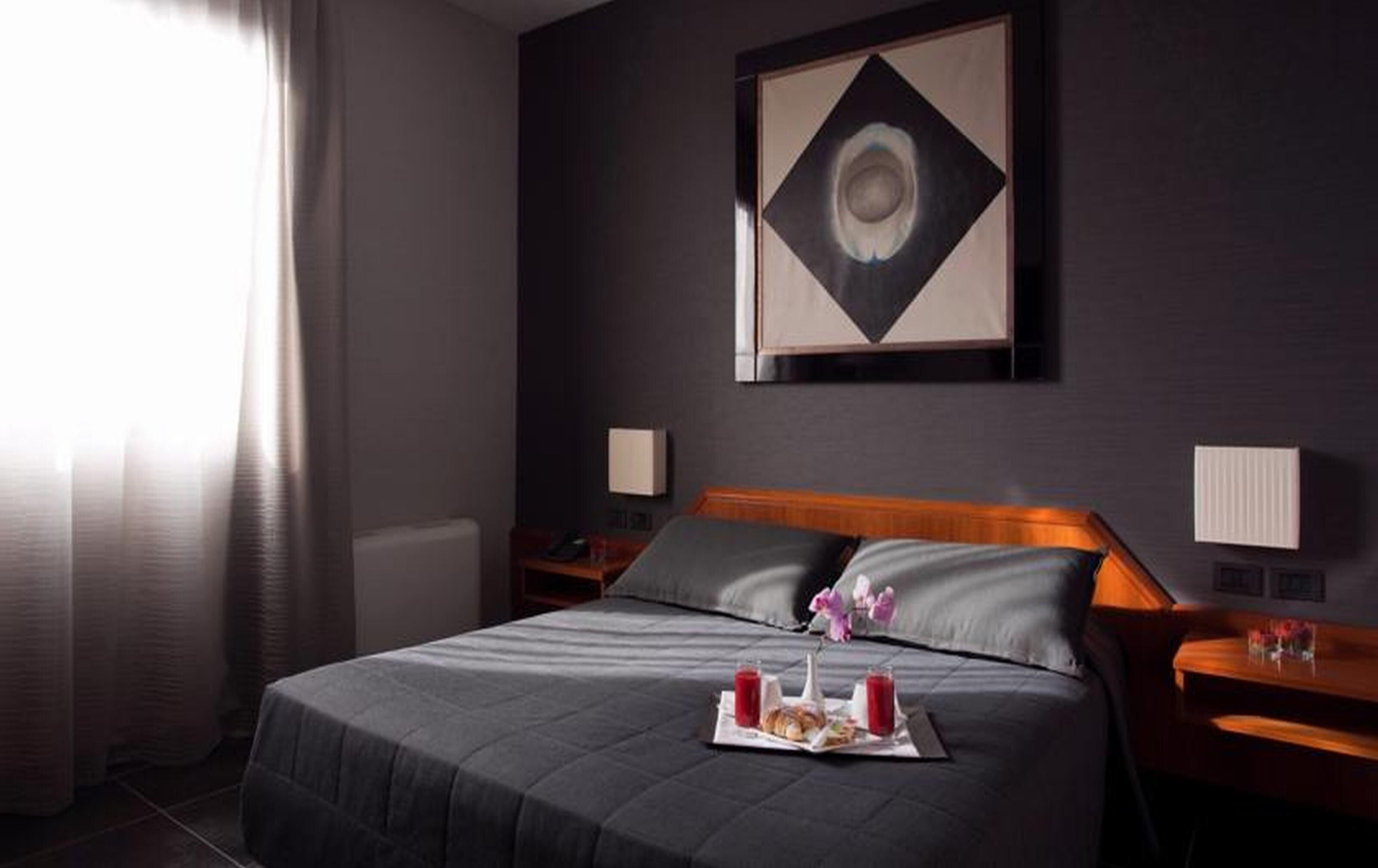 Una habitación de hotel con una cama grande, una mesita de noche con lámpara, una obra de arte en la pared y una ventana con cortinas.