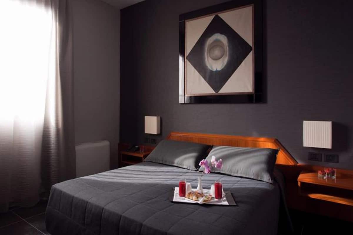 A hotel room with a large bed, a bedside table with a lamp, a wall-mounted artwork, and a window with curtains.