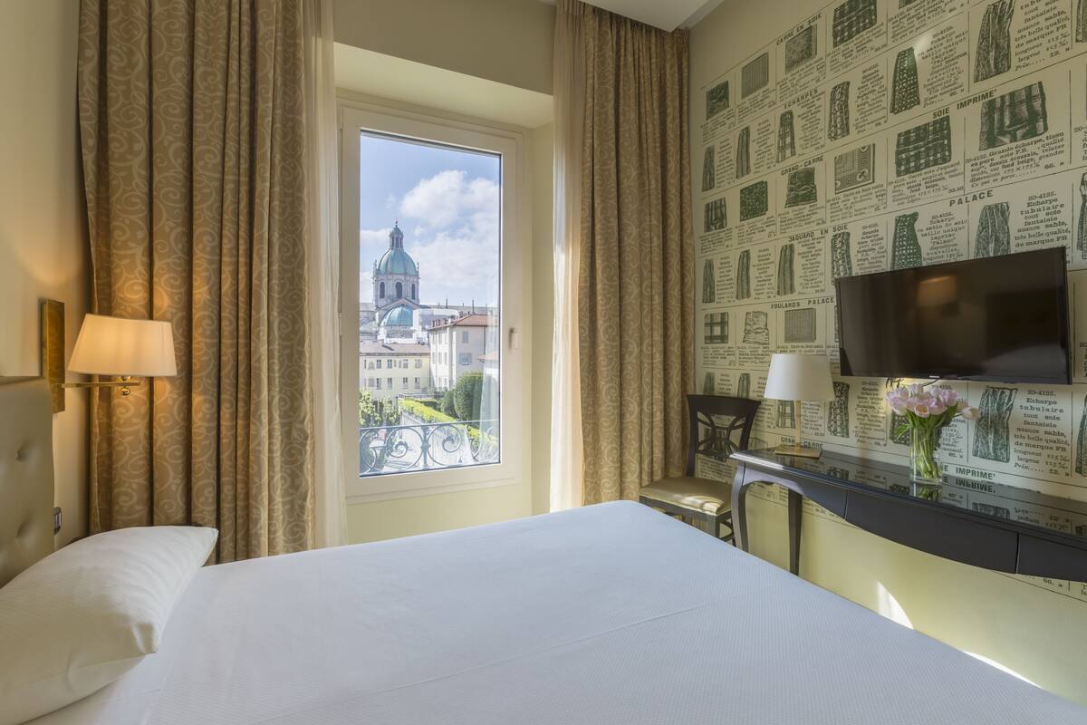 Une chambre d’hôtel avec un lit, un bureau avec une télévision, une fenêtre avec des rideaux et un mur orné de motifs imprimés.