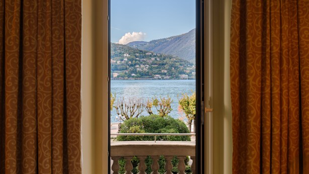 A view from a balcony with patterned curtains, overlooking a lake and a hillside with buildings. - Palace Hotel (Como)