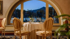 A dining area with a table set for two, a large arched window, and a view of a mountain. - Palace Hotel (Como)