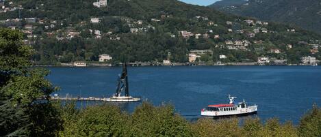 A lake with a boat and a statue in the foreground, surrounded by trees and a hillside with buildings in the background.