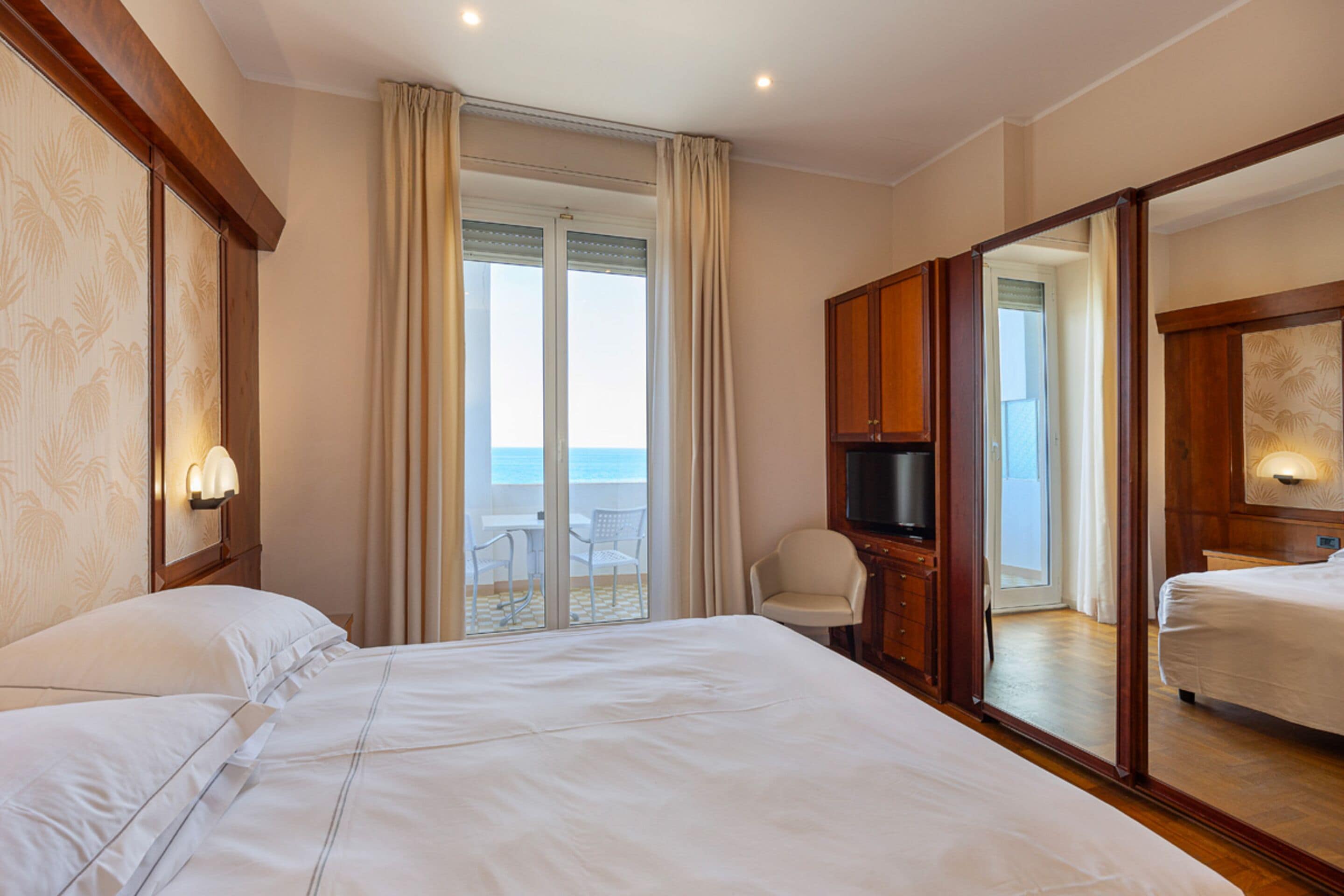 A hotel room with a large bed, a TV, a wardrobe, and a balcony with a view of the sea.
