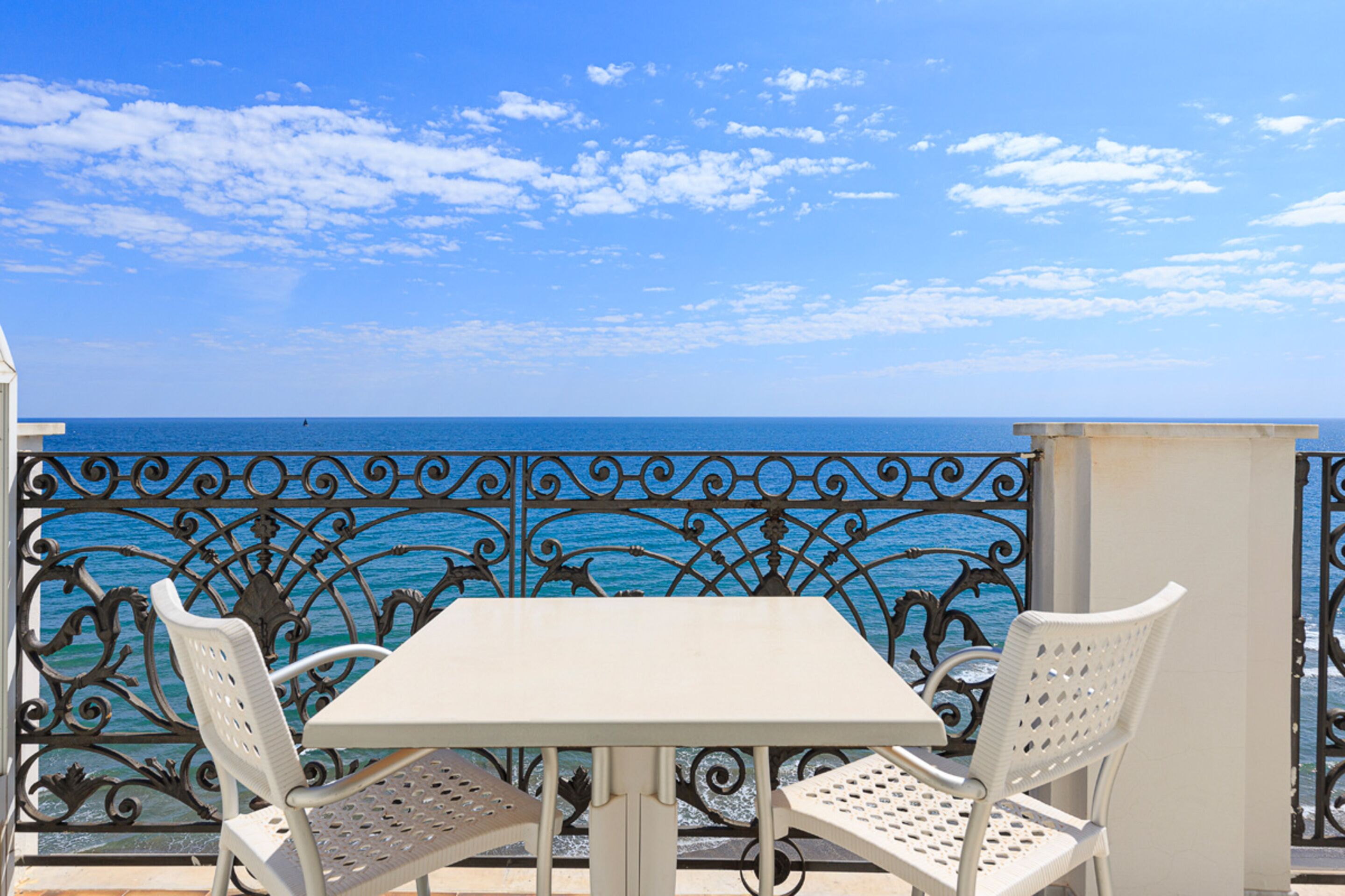 A balcony with a white table and two white chairs, overlooking a blue sea and sky.