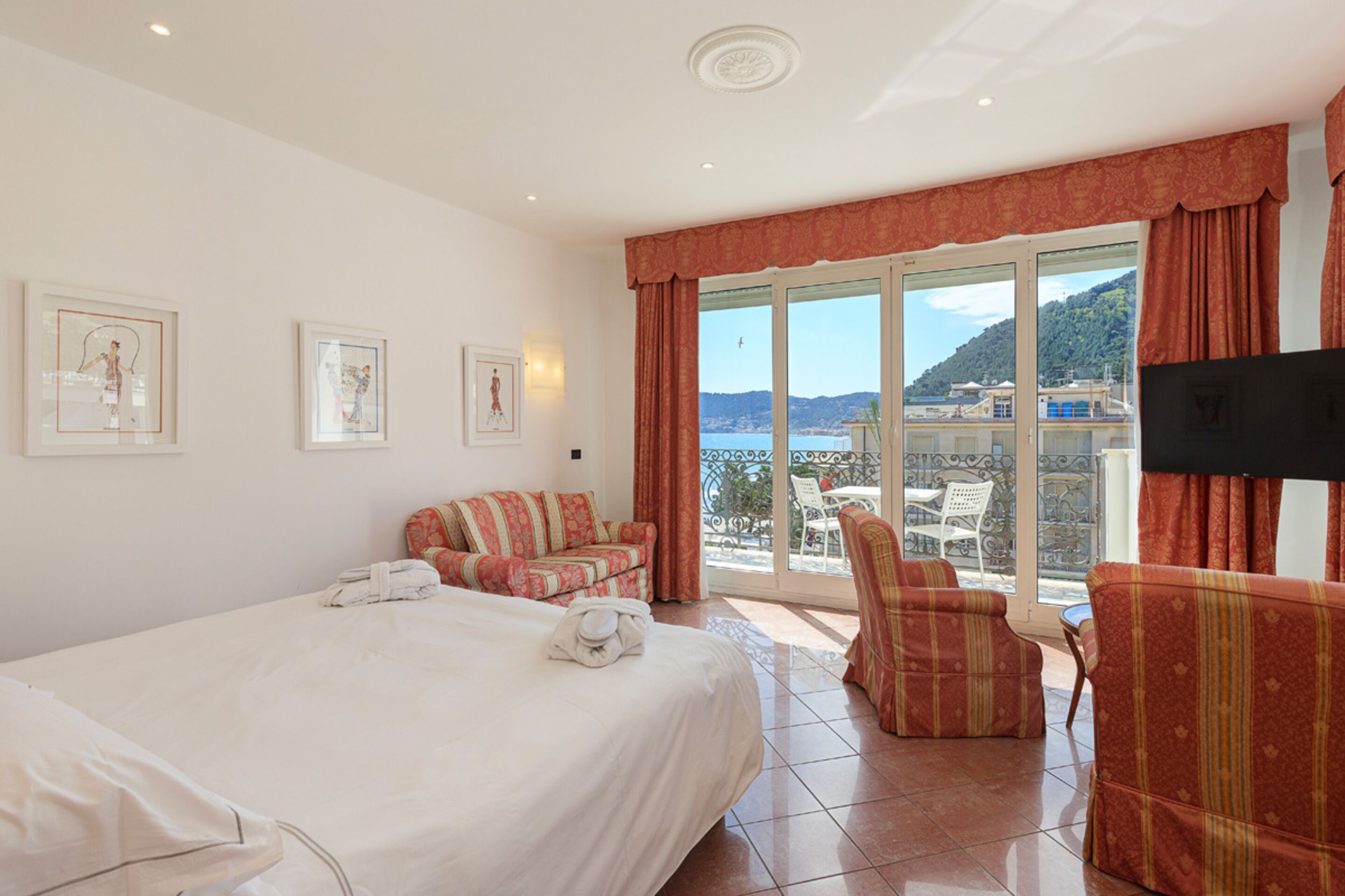 A hotel room with a bed, a sofa, a chair, and a TV. There are framed pictures on the wall and a view of the sea and hills outside the window.