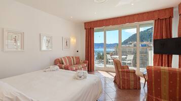 Ein Hotelzimmer mit Bett, Sofa, Sessel und Fernseher. An den Wänden hängen Bilder und durch das Fenster ist Meer und Hügellandschaft zu sehen.