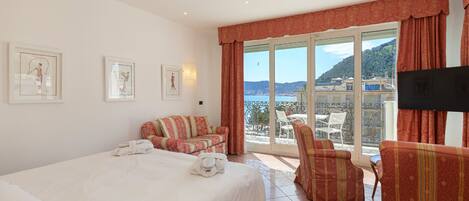 A hotel room with a bed, a sofa, a chair, and a TV. There are framed pictures on the wall and a view of the sea and hills outside the window.