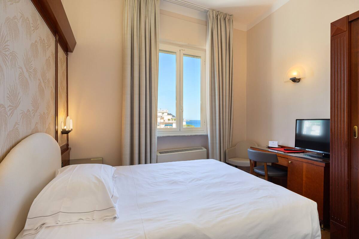 Ein Hotelzimmer mit einem Bett, einem Schreibtisch mit Computer, einem Fernseher und einem Fenster mit Vorhängen.