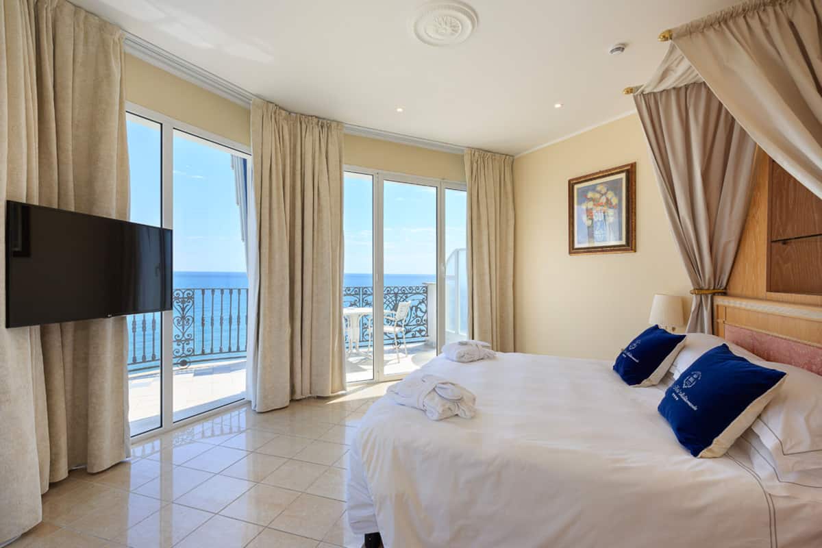 Ein Hotelzimmer mit Bett, Fernseher, Balkon mit Vorhängen und einem Bild an der Wand.