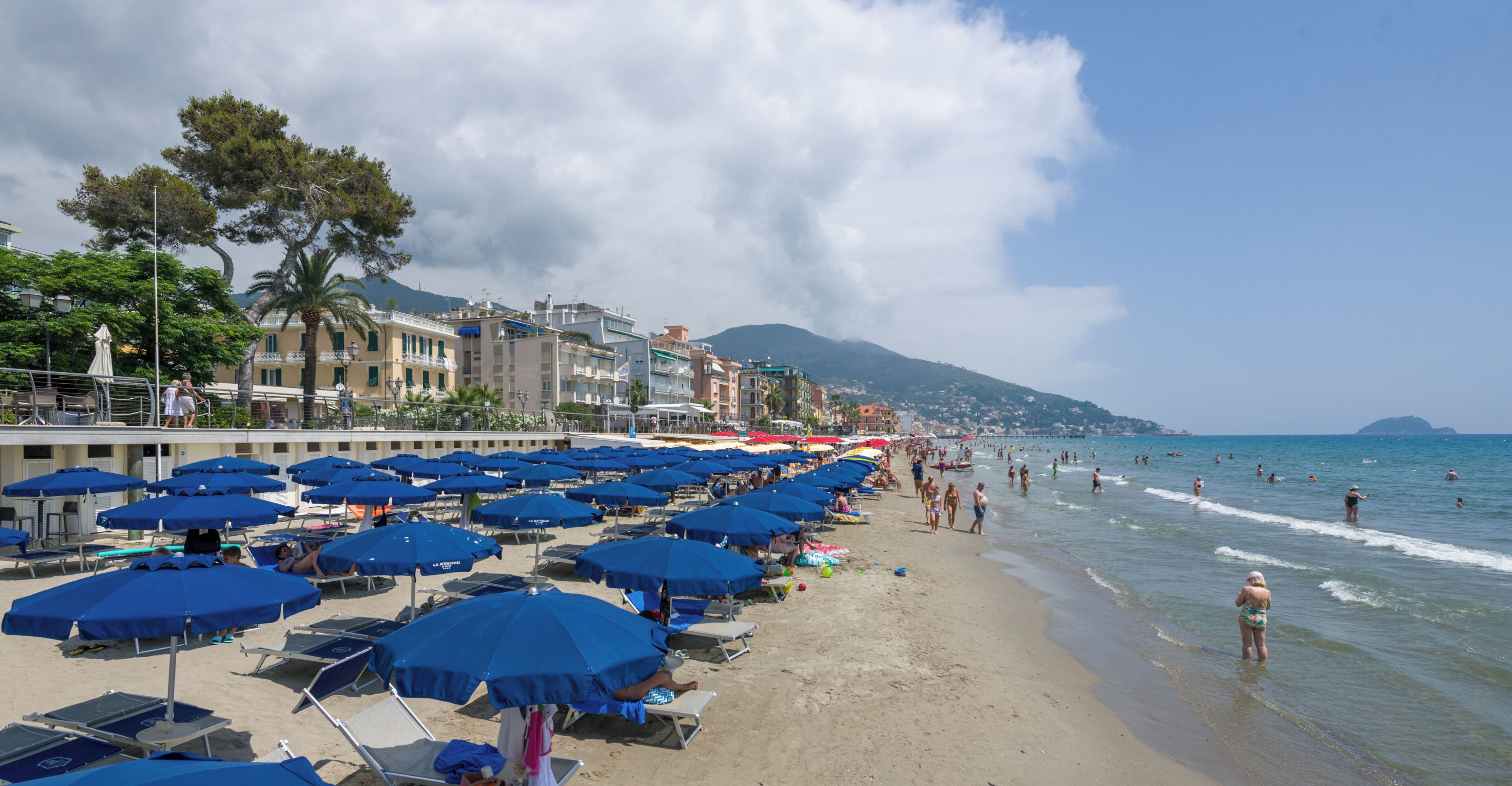 A beach with rows of blue umbrellas, a sandy shore, and buildings in the background.