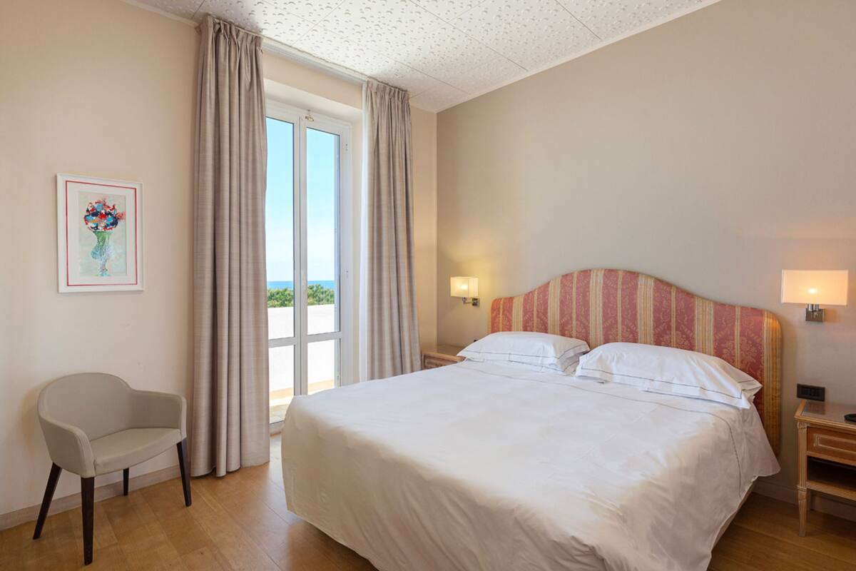 Ein Hotelzimmer mit Bett, Sessel, Nachttisch, Lampe und einem Fenster mit Vorhängen.