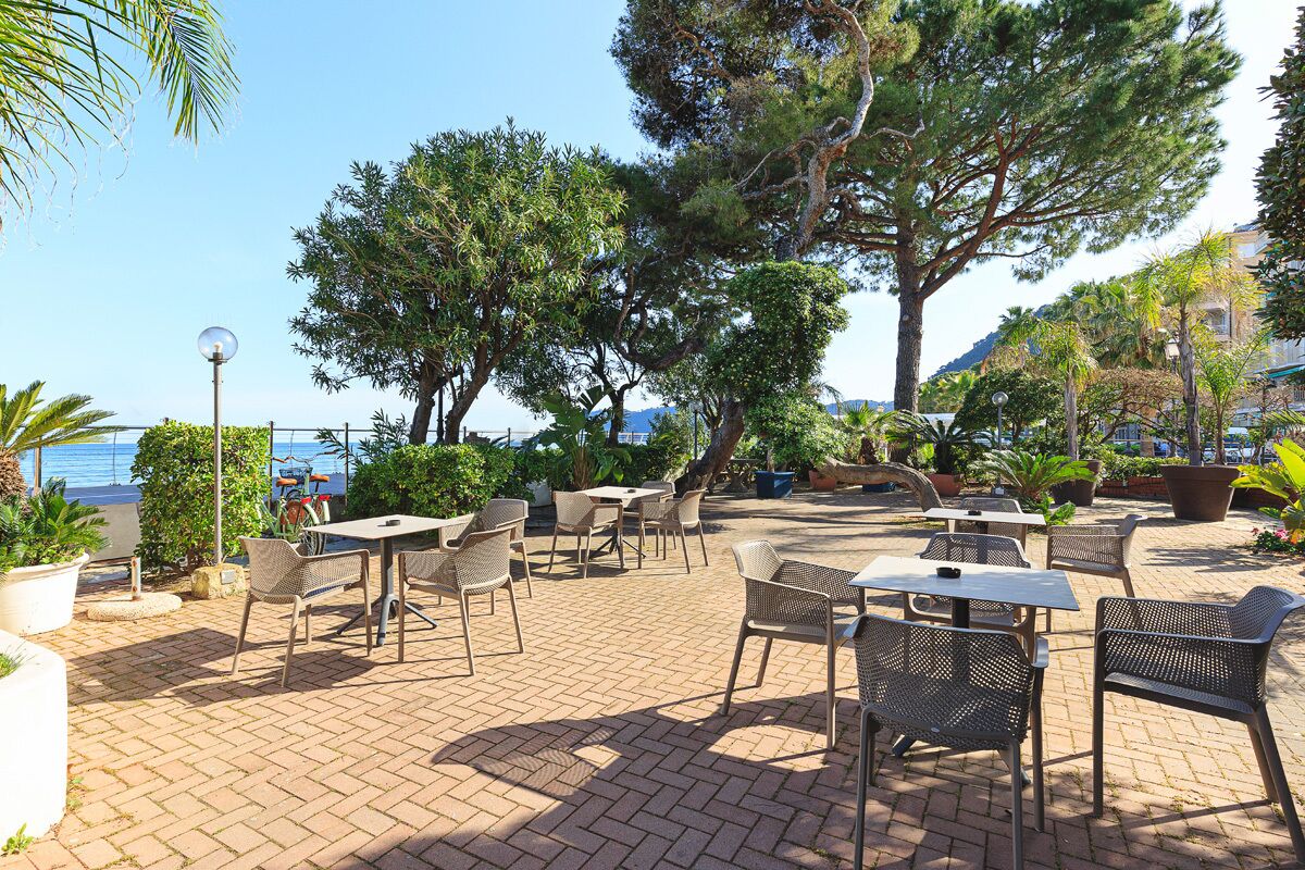 An outdoor patio area with tables and chairs, surrounded by trees and potted plants.